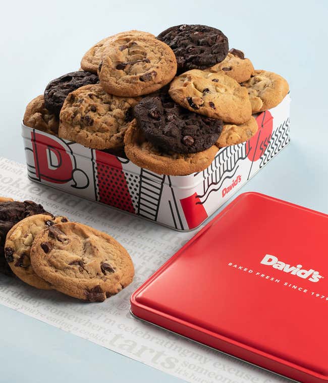  A decorative tin filled with an assortment of David's cookies, including chocolate chip, double chocolate, and oatmeal raisin. The tin features a colorful red, white, and black design with the David's logo. A few cookies and the tin's red lid are placed 