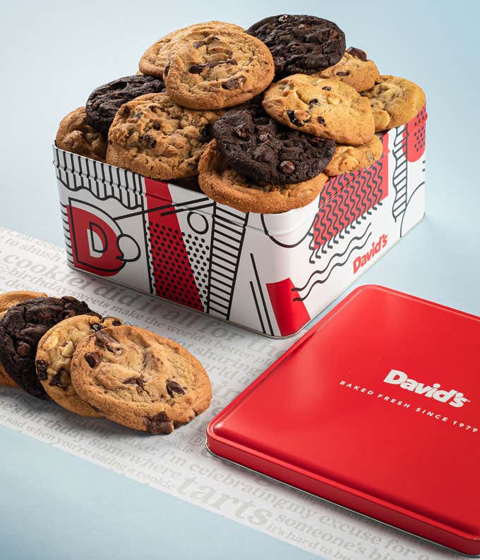  A decorative tin filled with David's cookies, including chocolate chip, double chocolate, and oatmeal raisin. The tin features a red, white, and black design with the David's logo. A few cookies and the red lid are placed beside it.