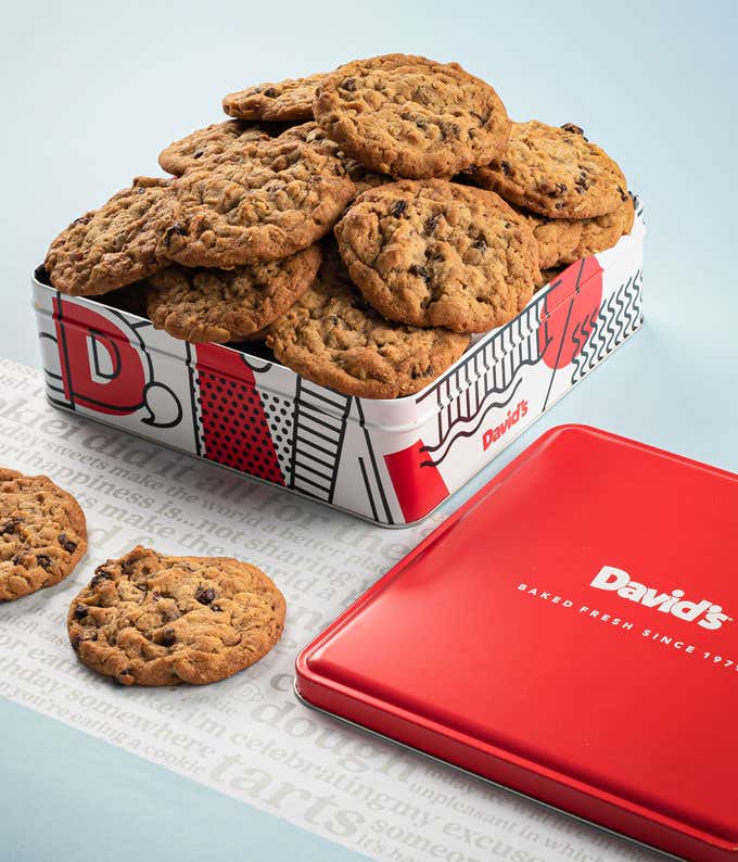 A decorative tin filled with David's oatmeal raisin cookies. The tin features a red, white, and black design with the David's logo. A few cookies and the red lid are placed beside the tin.