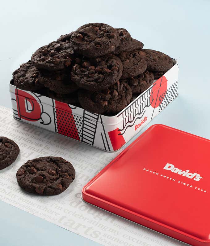  A decorative tin filled with David's double chocolate cookies. The tin features a red, white, and black design with the David's logo. A few cookies and the red lid are placed beside the tin.