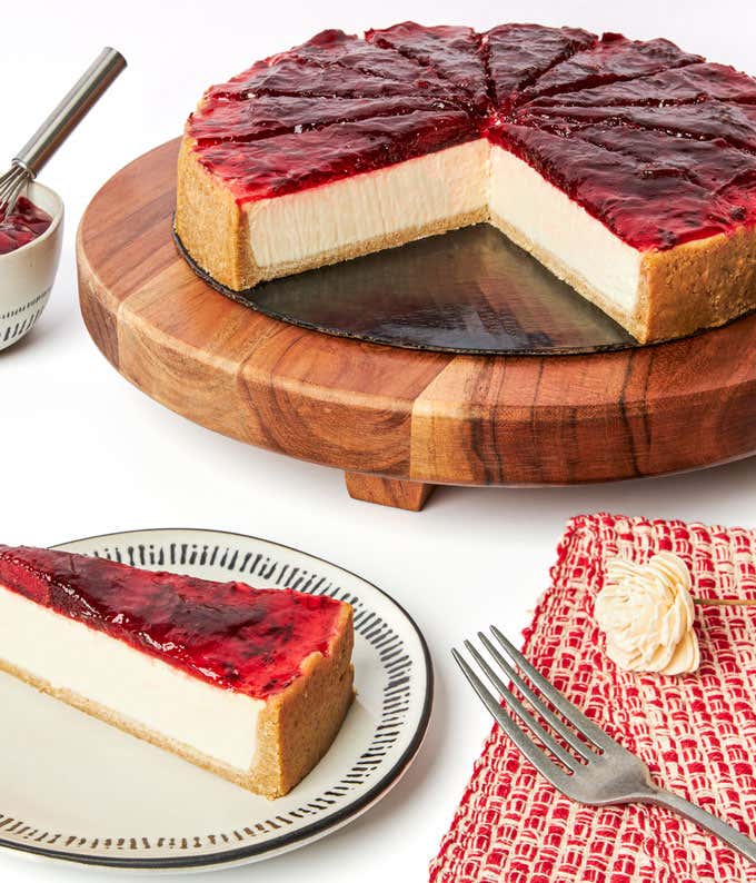 A strawberry cheesecake on a wooden cake stand, with a slice cut out and placed on a plate beside it. The cheesecake features a thick layer of strawberry topping and a graham cracker crust. A bowl of extra strawberry topping and a fork are also visible.