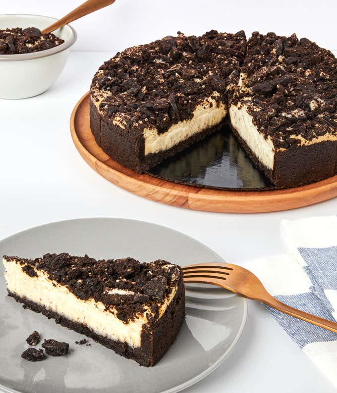 A cookies and cream cheesecake on a wooden cake stand, with a slice on a plate beside it. The cheesecake has a creamy filling, chocolate cookie crust, and crushed cookies on top. A bowl of extra cookie crumbs and a fork are also visible.