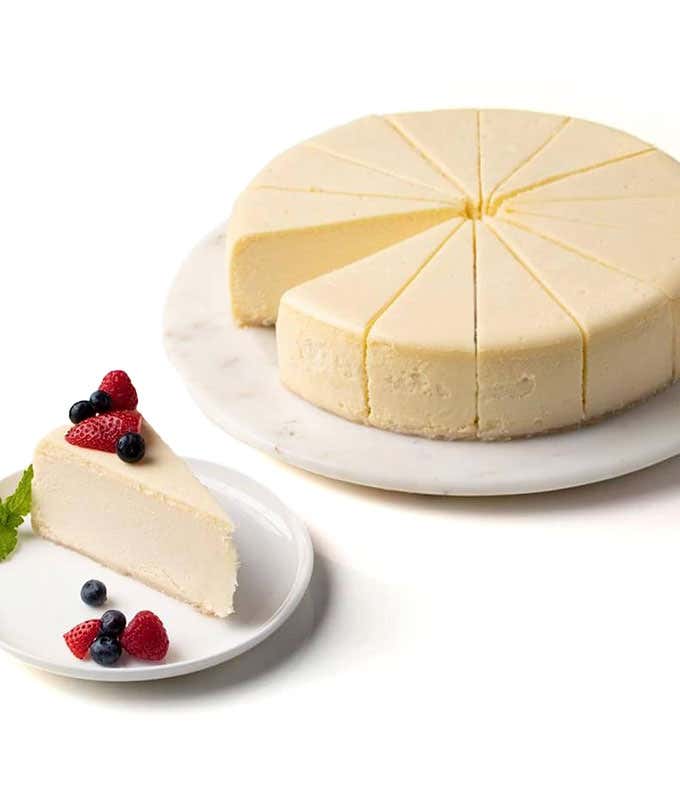 A sugar-free New York cheesecake on a marble platter, with a slice on a plate beside it. The slice is garnished with fresh berries and a mint leaf. The cheesecake is creamy and smooth, with cleanly cut slices.