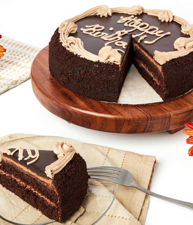 A chocolate birthday cake with &quot;Happy Birthday&quot; written on top in frosting, displayed on a wooden cake stand. A slice is cut out and placed on a plate beside the cake. The cake has chocolate layers and frosting decorations.