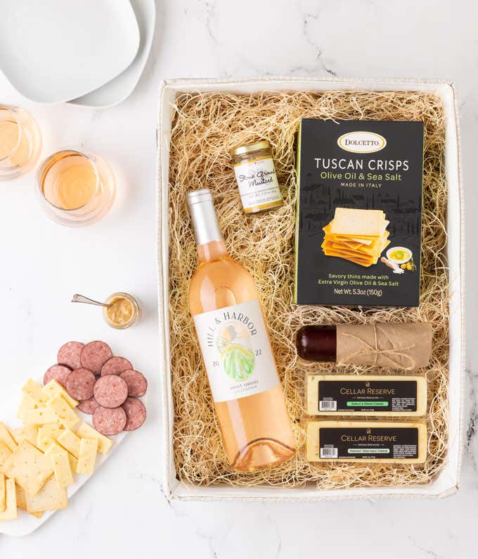 A gift box containing Tuscan crisps, salami, cheeses, mustard, and a bottle of Hill & Harbor ros wine. Surrounding the box are plates with sliced cheese and salami, mustard, and two glasses of wine. 