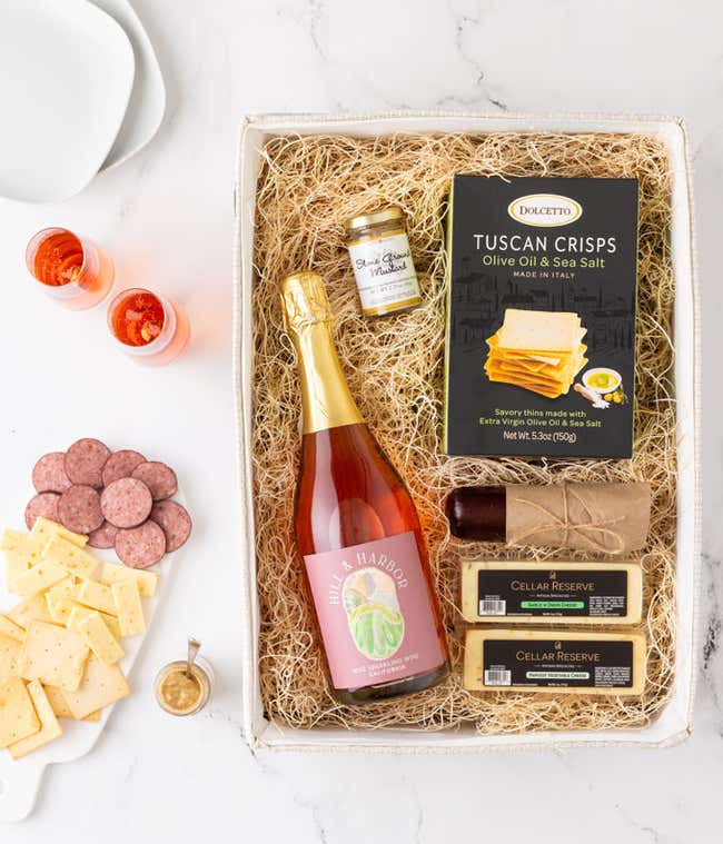 A gift box with Tuscan crisps, salami, cheeses, mustard, and a bottle of Hill &amp; Harbor sparkling wine. Surrounding the box are plates with cheese, salami, mustard, and two glasses of sparkling wine.