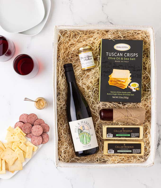 a gift basket with contents laid out across a white wood table. A bottle of red wine, a glass of red wine, a block of cheddar cheese, a summer sausage, a jar of honey mustard, a box of crackers, with a cutting board of sliced meat and cheese 