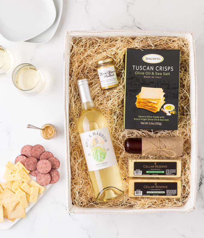 a gift basket with contents laid out across a white wood table. A bottle of white wine, a glass of white wine, a block of cheddar cheese, a summer sausage, a jar of honey mustard, a box of crackers, with a cutting board of sliced meat and cheese 