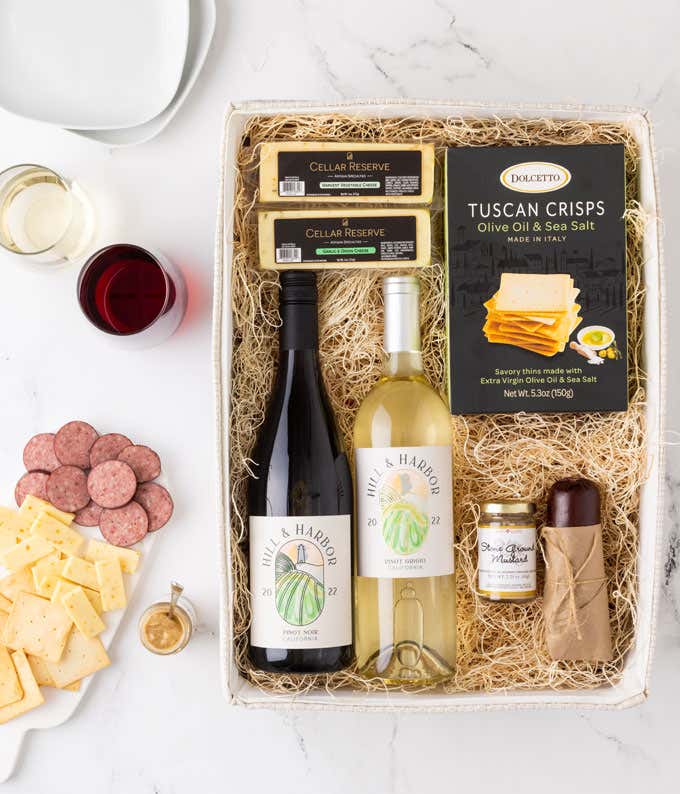 a gift basket with contents laid out across a white wood table. A bottle of red wine, a glass of red wine, a bottle of white wine, a glass of white wine, a block of cheddar cheese, a summer sausage, a jar of honey mustard, a box of crackers, with a cuttin