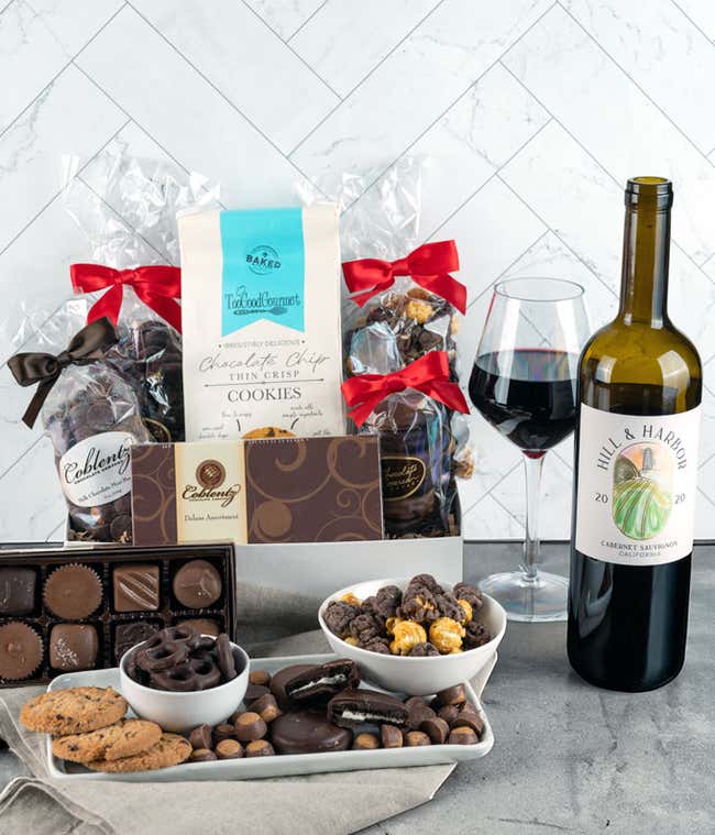 Gourmet gift set featuring assorted chocolates, chocolate chip cookies, a bottle of Hill &amp; Harbor Cabernet Sauvignon, and a glass of red wine, displayed on a table with festive packaging.