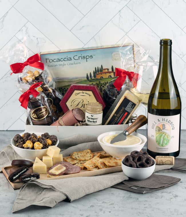 Gourmet gift box featuring focaccia crisps, brie cheese, cheddar cheese, summer sausage, mustard, chocolate-covered snacks, and a bottle of Hill &amp; Harbor Chardonnay, with a glass of white wine and an assortment of crackers, cheese, and sausage displayed o