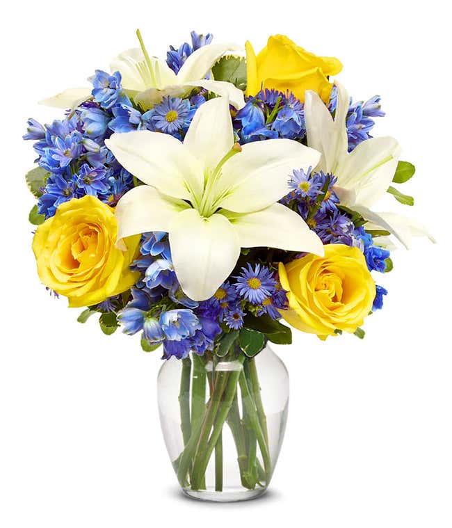 An elegant floral arrangement featuring white Asiatic lilies, yellow roses, blue delphinium, and purple Monte Casino, optionally arranged in a glass vase. This vibrant mix offers a stunning and colorful display.