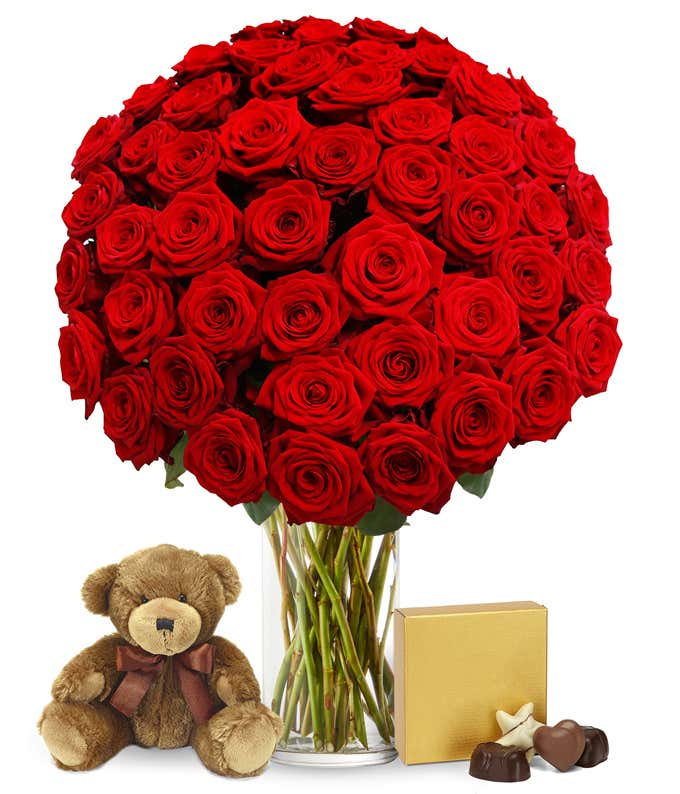 Image of 50 long-stemmed red roses in a tall cylinder vase, accompanied by a box of chocolates and a cuddly teddy bear, the perfect gift for expressing love and affection.