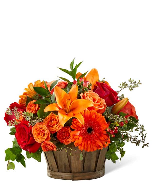 A vibrant floral arrangement featuring red roses, orange roses, orange spray roses, orange gerbera daisies, orange Asiatic lilies, red hypericum berries, and lush greens, all beautifully presented in a wooden basket.