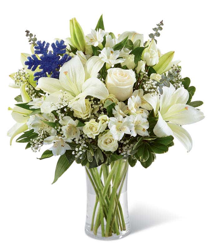 White lilies, white roses (Deluxe and Premium only), white spray roses, white alstroemeria, baby's breath, and blue snowflake in a keepsake vase, creating a serene and memorable floral arrangement.