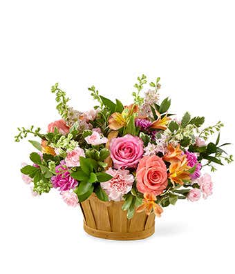 A floral arrangement featuring pink and coral roses (deluxe and premium sizes), pink carnations, pink mini carnations, orange alstroemeria, and pink larkspur, arranged in a wooden basket.