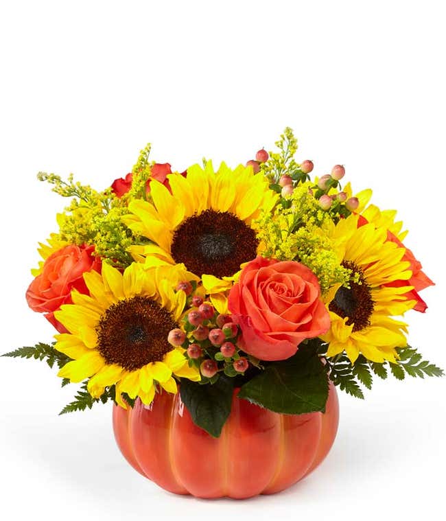 A cheerful floral arrangement featuring sunflowers, orange roses, yellow solidago, and peach berries, all beautifully arranged in a pumpkin container.