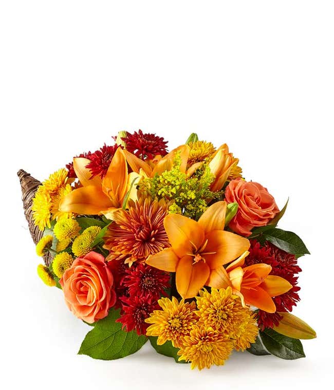 A festive arrangement featuring orange lilies, solidago, red cushion poms, butterscotch cushion poms, yellow button poms, deluxe and premium orange roses, deluxe and premium butterscotch mums, and seasonal greens, all beautifully displayed in a cornucopia