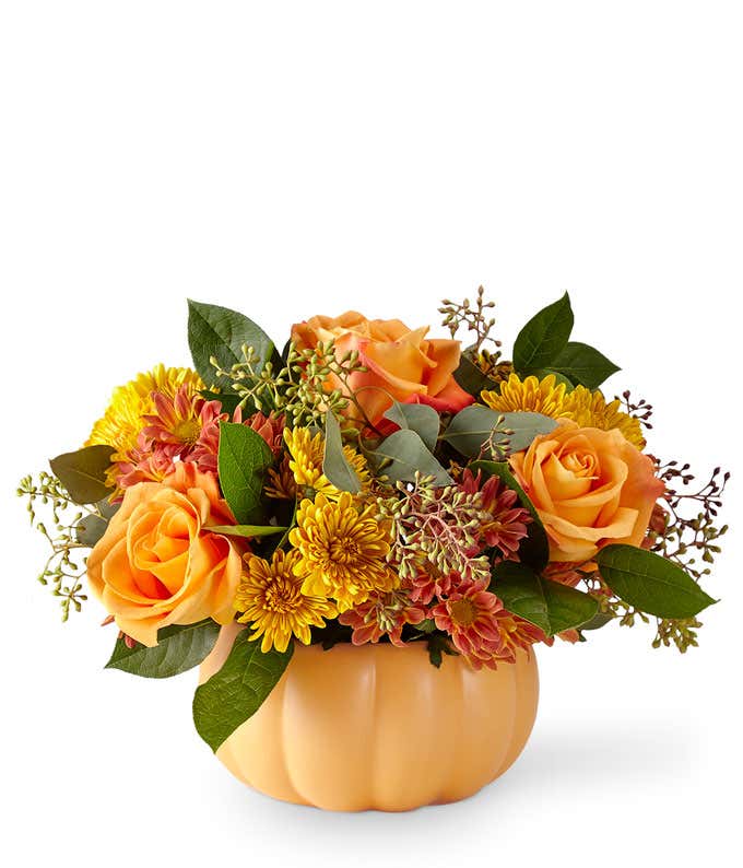  A warm and festive floral arrangement featuring butterscotch cushion poms, bronze daisies, orange roses, and seasonal greens, all beautifully presented in a ceramic pumpkin.