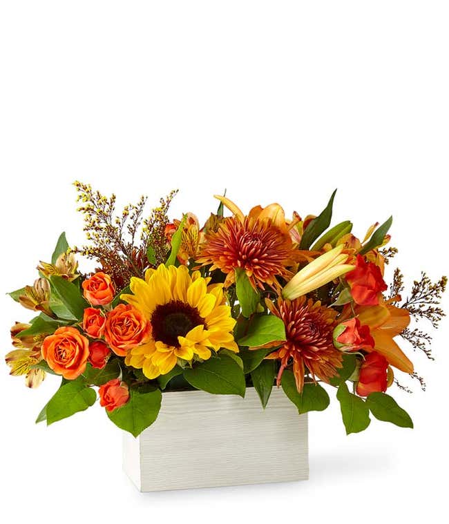 A vibrant floral arrangement featuring orange spray roses, yellow alstroemeria, butterscotch mums, red-tinted solidago, sunflowers, deluxe and premium orange lilies, and seasonal greens, all beautifully arranged in a white wooden box.