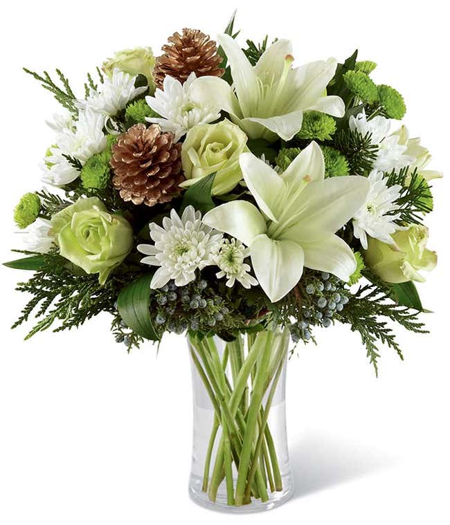 A festive arrangement with white Asiatic lilies, green roses, green and white poms, evergreens, displayed in a glass cylinder vase.