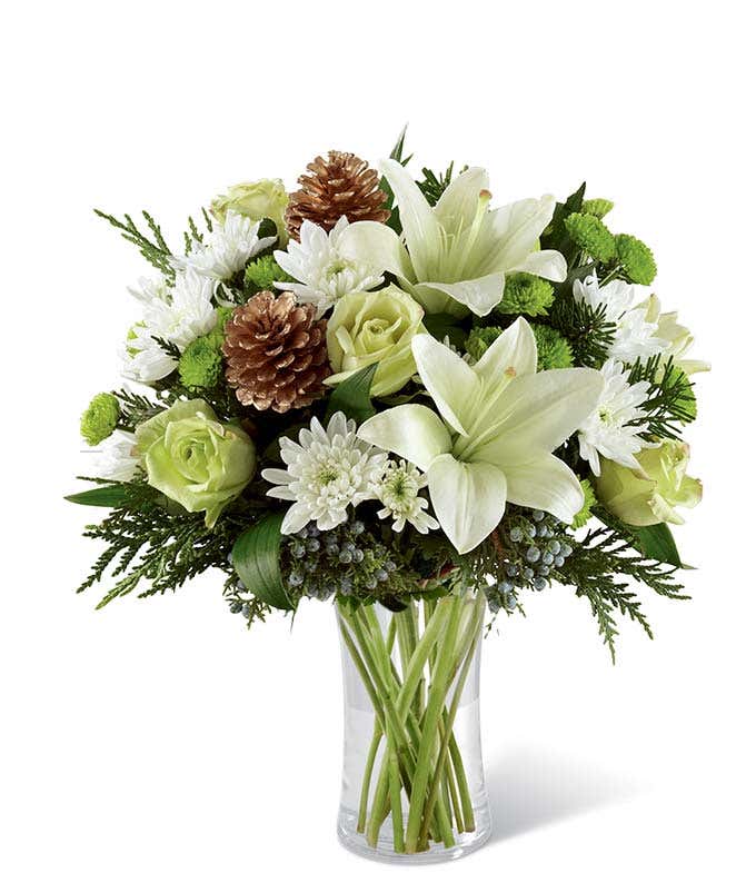 A festive arrangement with white Asiatic lilies, green roses, green and white poms, evergreens, displayed in a glass cylinder vase.