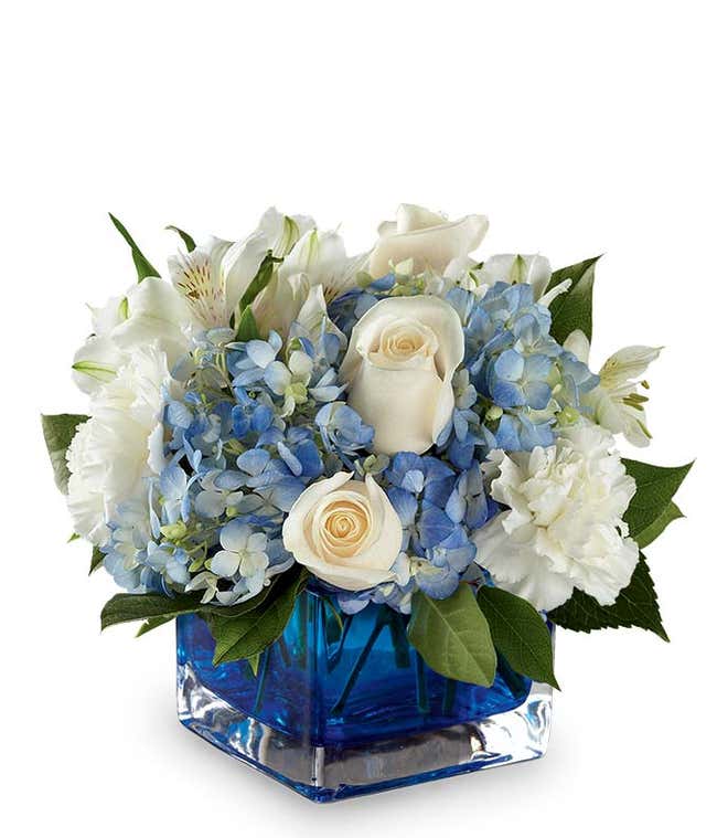 A serene arrangement featuring white carnations, white roses, white alstroemeria, and blue hydrangea, elegantly presented in a blue cube vase. This beautiful mix creates a peaceful and soothing display.