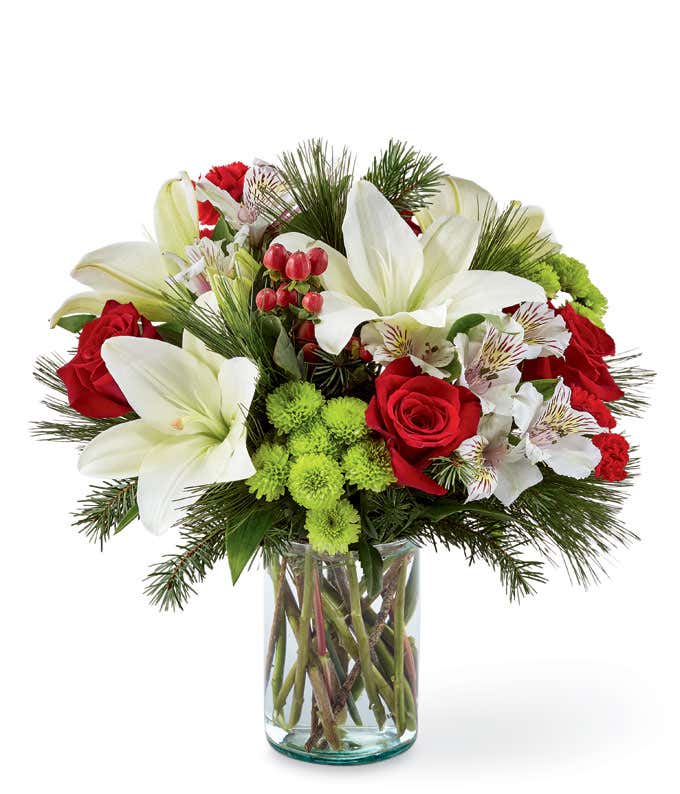 A festive arrangement with white lilies, white alstroemeria, red roses, red mini carnations, evergreens, displayed in a clear vase.