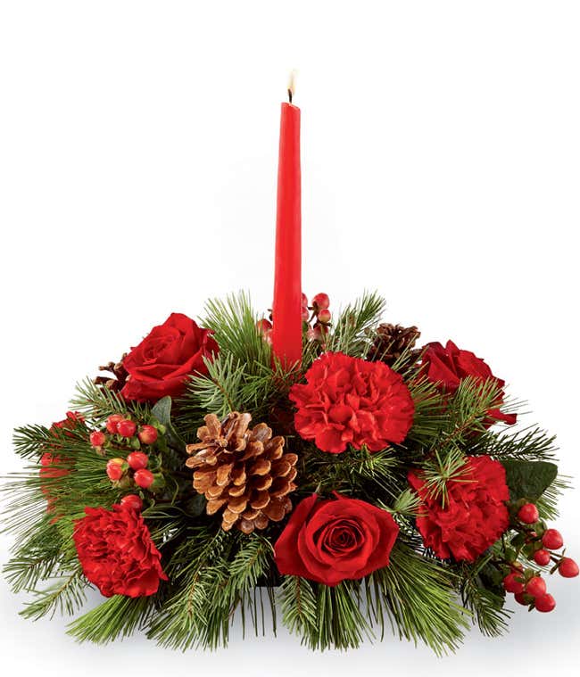 Festive centerpiece with a red candle in the middle, surrounded by red roses, red carnations, pine cones, red berries, and evergreen branches, creating a classic holiday display.