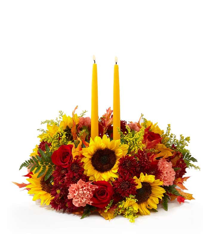  A warm and vibrant floral arrangement featuring sunflowers, red roses, orange carnations, red daisies, bronze daisies, and oak leaves, complemented by golden yellow candles.