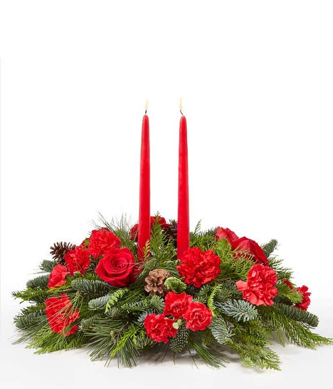 A festive arrangement with red roses, red carnations, red mini carnations, assorted seasonal greens, and red taper candles.