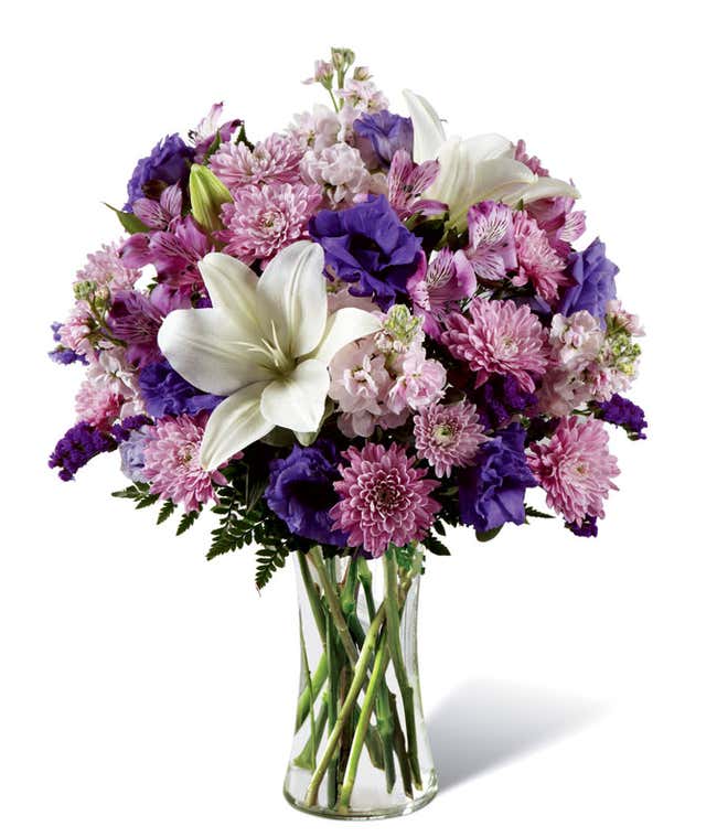 Floral arrangement featuring white Asiatic lilies, lavender stock, purple alstroemeria, purple lisianthus, lavender cushion poms, purple statice, leatherleaf fern, in a tall cylinder vase.