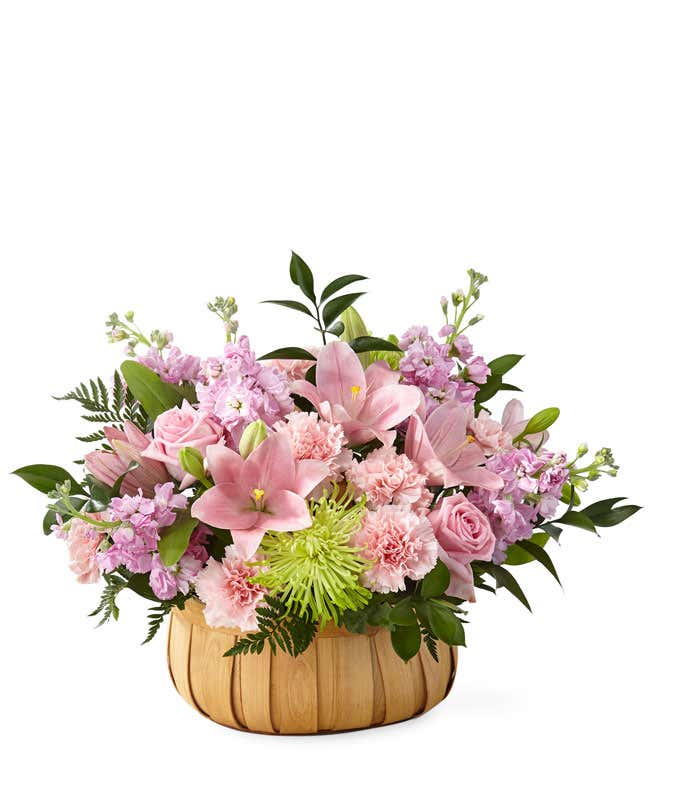 Pink lilies, roes, and carnations, with green poms and fresh floral greens arranged into a wooden basket