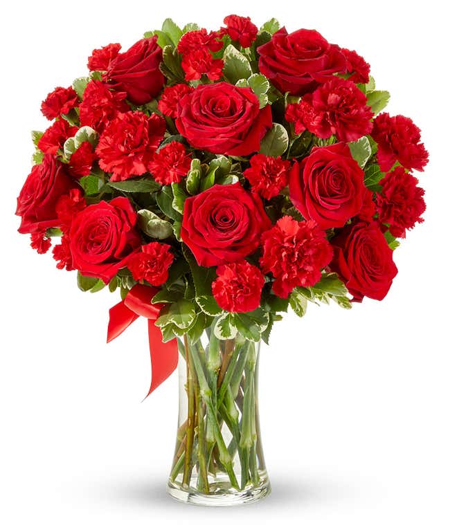 Floral arrangement featuring red roses, red carnations, red mini carnations, and a decorative red ribbon, florist delivered in a clear glass vase.