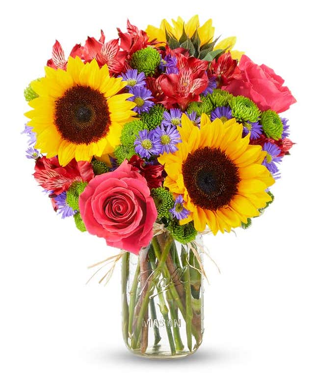 Floral arrangement featuring sunflowers, red alstroemeria, pink roses (in deluxe and premium versions), purple monte casino, and green button poms in a mason jar vase, with a personalized card message.