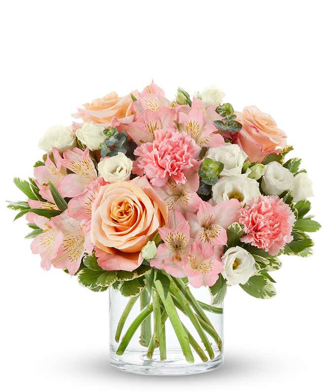 A delightful floral arrangement of peach roses, pink carnations, pink alstroemeria, and white lisianthus, elegantly arranged in a glass vase. This soft and romantic mix of flowers offers a gentle and inviting display.