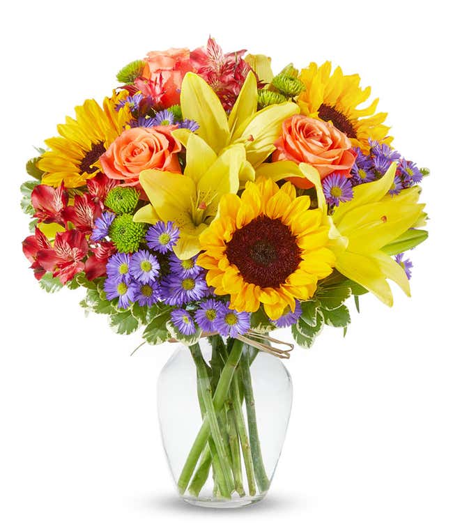 A radiant floral arrangement featuring sunflowers, yellow lilies, orange roses, alstroemeria, Monte Casino, and green poms, all beautifully arranged in a clear glass vase. This vibrant mix creates a warm and inviting display.