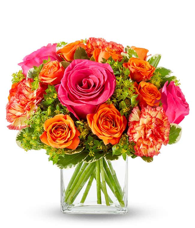 A lively floral arrangement featuring hot pink roses, orange spray roses, orange carnations, bupleurum, lily grass, and pittosporum, all beautifully arranged in a clear glass cube vase. This vibrant mix creates a stunning and energetic display.