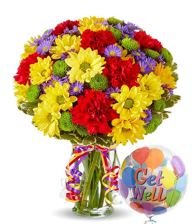 A cheerful floral arrangement featuring red carnations, yellow daisies, purple Monte Casino, and green button poms in a vase decorated with rainbow ribbons, accompanied by a 'Get Well' mylar balloon.