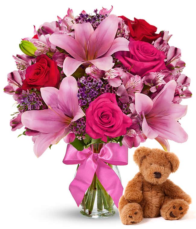 Red and pink roses, pink alstroemeria in a clear vase with bow, accompanied by a keepsake teddy bear.