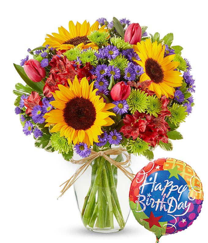 a colorful bouquet in a clear vase, featuring bright yellow sunflowers, red tulips, purple asters, and lush green foliage. The arrangement is tied with a rustic twine bow. Next to the flowers there's a 'Happy Birthday' mylar balloon.