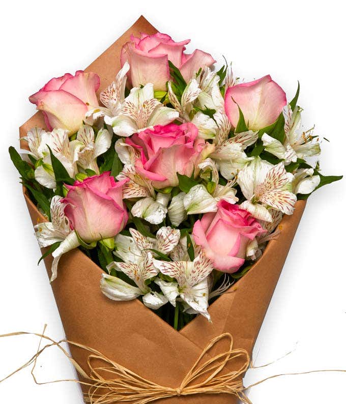 A bouquet of pink roses and white alstroemeria with fresh greens, wrapped in brown parchment paper and tied with twine.