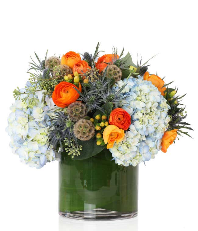  A stunning floral arrangement featuring blue hydrangea, berries, orange ranunculus, and eucalyptus, all elegantly presented in a cylinder glass vase.