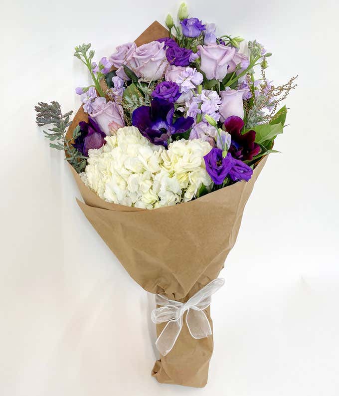  Bouquet of light purple roses, dark purple flowers, and white hydrangeas wrapped in brown paper and tied with a white ribbon.