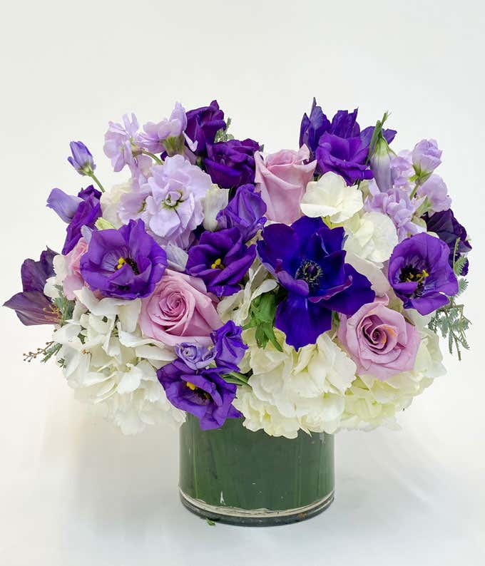 Elegant bouquet featuring a mix of purple and lavender flowers, including roses and lisianthus, accented with white hydrangeas, arranged in a clear glass vase for a sophisticated and beautiful display.