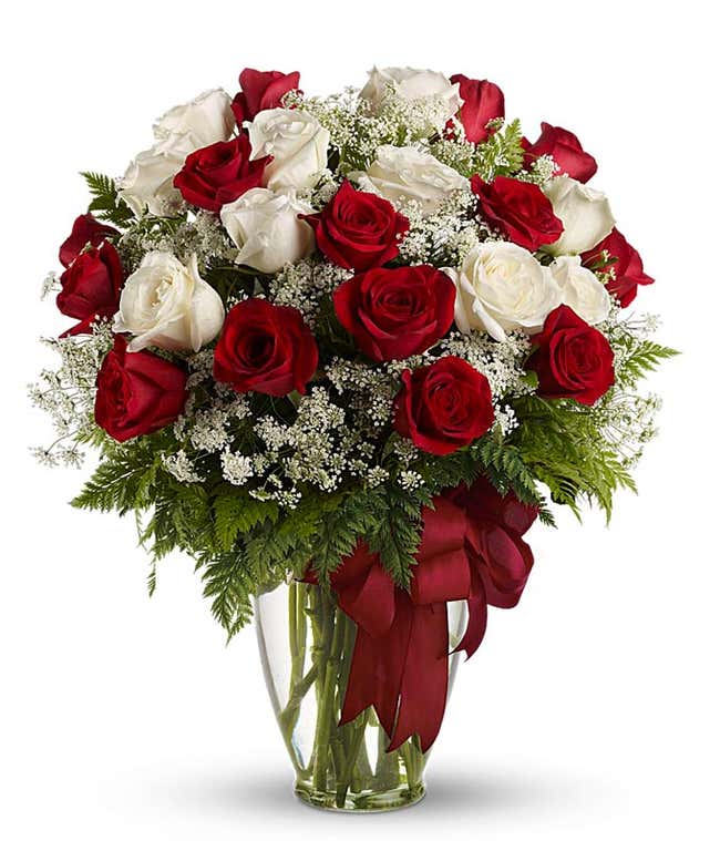 A floral arrangement featuring red roses, white roses, and floral greens in a clear glass vase with a red bow.