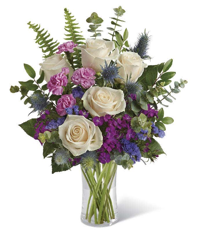 A floral arrangement in a glass cylinder vase featuring crme roses, pink mini carnations, burgundy Sweet William, purple sinuata statice, blue eryngium, huckleberry, spiral eucalyptus, and sword fern.