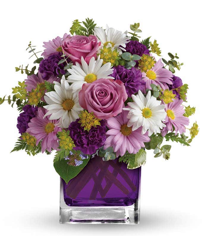 A beautiful arrangement featuring purple carnations, lavender daisy spray mums, white daisy spray mums, bupleurum, spiral eucalyptus, and leatherleaf fern, presented in a purple glass cube vase. Deluxe and premium bouquets include purple roses for an extr