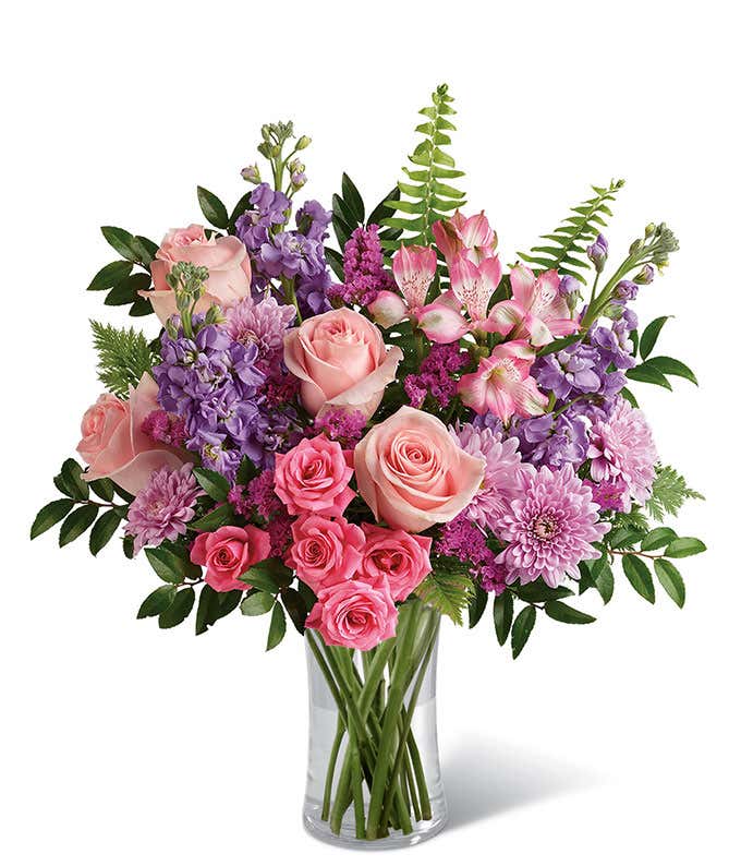 Floral arrangement featuring light pink roses, pink spray roses, pink alstroemeria, lavender stock, lavender mums, raspberry statice, and greenery in a glass vase.