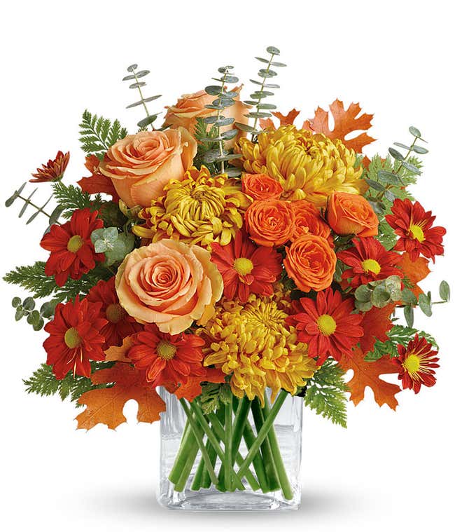 A stunning floral arrangement featuring deluxe and premium orange roses, orange spray roses, bronze mums, and red daisies, accented with spiral eucalyptus, all elegantly displayed in a clear cube vase.